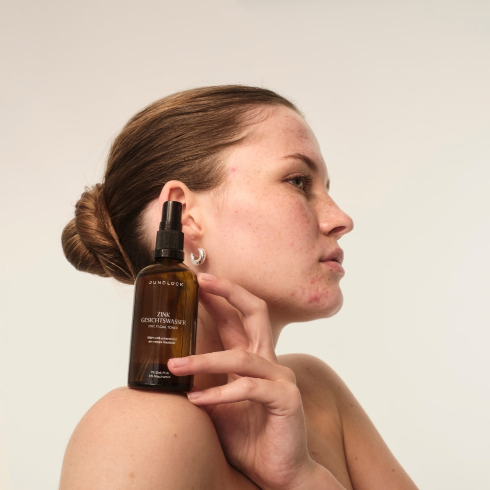 Eine Frau hält eine Braunglasflasche mit dem Produkt "ZINK GESICHTSWASSER" in der Hand. Ihr Gesicht zeigt natürliche