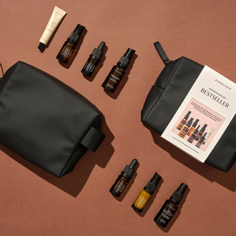 Black zippered cosmetic bag with eight amber and beige skincare bottles and tubes on brown background, including a booklet.