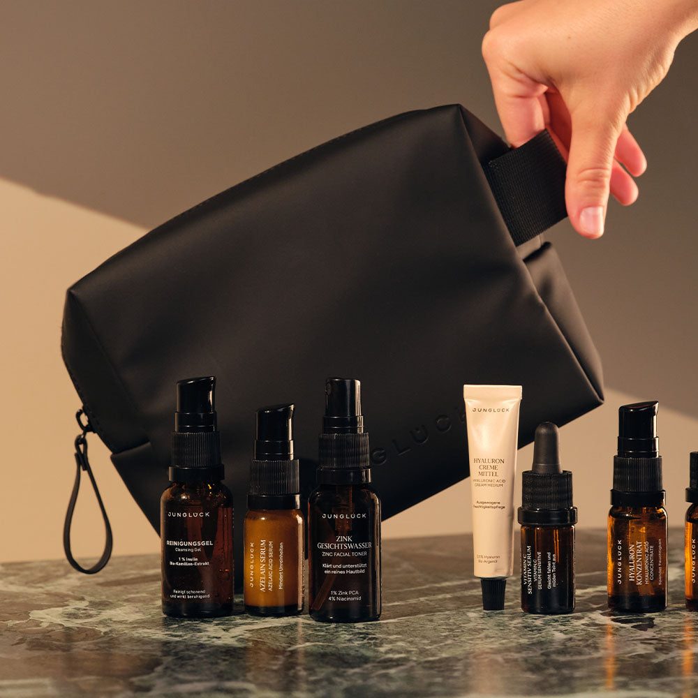Black cosmetic bag with four amber dropper bottles, a small beige tube, and a spray bottle on a marble surface.