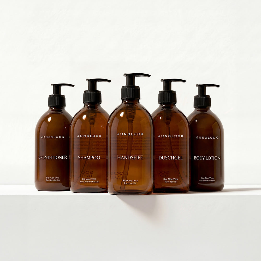 Fünf Braunglasflaschen mit Pumpspendern: Conditioner, Shampoo, Handseife, Duschgel und Body Lotion, alle mit weißen